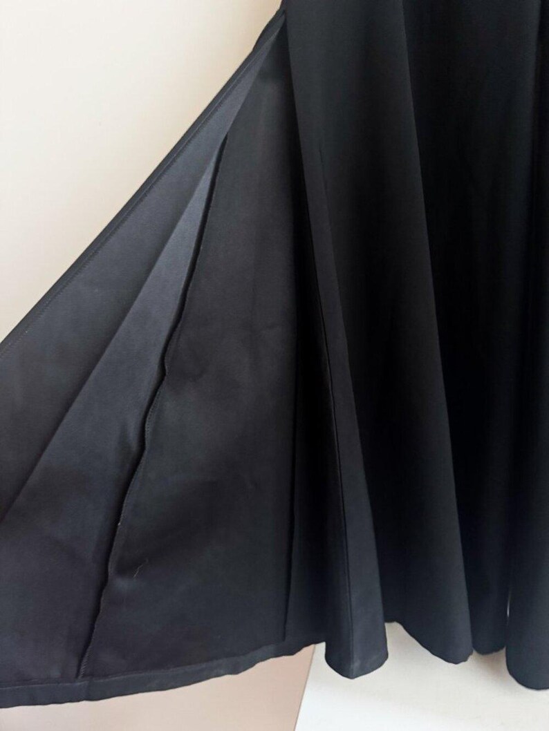 May include: A black skirt featuring a deep side slit that reveals the inner lining of the fabric. The garment is constructed with multiple vertical panels that create a pleated, flowing silhouette, extending downward in a full, flared shape.