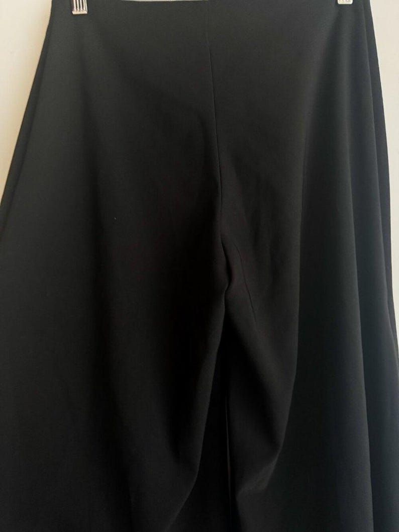 May include: A pair of solid black wide-leg trousers featuring a high-waisted design with a central vertical seam running down the front. The garment is constructed from a smooth, opaque fabric and is suspended from a metal hanger at the top.