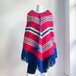 May include: A red, white, and blue crocheted poncho with blue fringe. The poncho has a geometric pattern and a pom-pom detail.