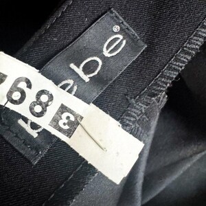 May include: A black garment featuring a black fabric label with the white text bebe stitched onto the interior. A white paper tag with the black printed number 389 is attached to the garment with a thin metal staple, partially obscuring the label.