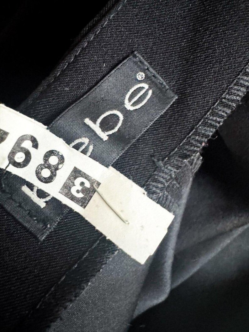 May include: A black garment featuring a black fabric label with the white text bebe stitched onto the interior. A white paper tag with the black printed number 389 is attached to the garment with a thin metal staple, partially obscuring the label.