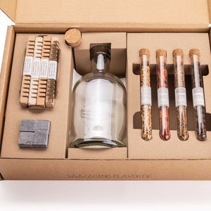 Peut inclure: Une boîte en carton marron contenant une bouteille en verre transparent, quatre tubes à essai en verre avec bouchons en liège et une pile de bâtonnets en bois. Les tubes à essai contiennent des épices et la boîte porte l'inscription "WWW.AGING-FLAVOR.DE" imprimée au bas.