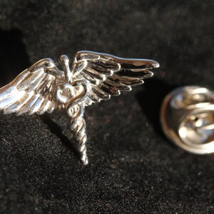 May include: Silver-toned winged medical caduceus pin with a safety clasp. The wings are spread wide and the staff is coiled with two snakes.
