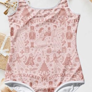 May include: A pink one-piece swimsuit featuring a repeating pattern of Nutcracker characters, Christmas trees, and snowflakes. The swimsuit has a white trim and the word "NUTCRACKER" is printed on the front.