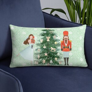May include: A green and white throw pillow with a Christmas tree, a ballerina, and a nutcracker. The tree is decorated with ornaments, a star, and a rocking horse. The ballerina is wearing a blue dress and holding a doll. The nutcracker is wearing a red and blue uniform.