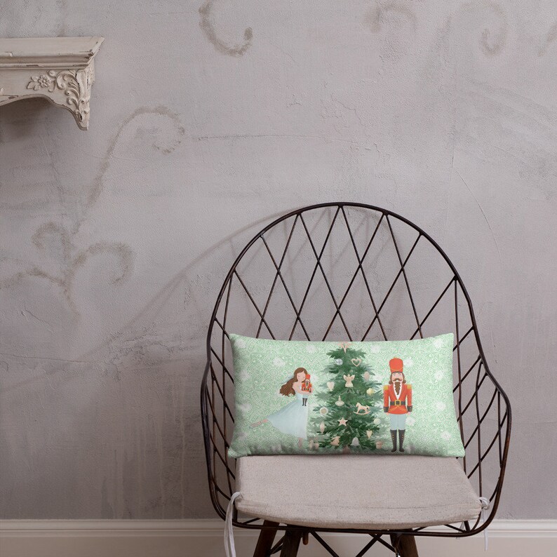 May include: A green and white throw pillow with a Christmas scene featuring a girl in a blue dress, a nutcracker, and a decorated Christmas tree.