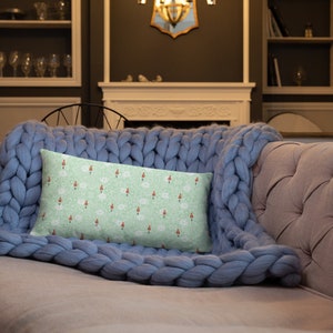 May include: A light green throw pillow with a repeating pattern of red and white figures on a light green background. The pillow is on a blue knitted throw blanket on a light gray couch.