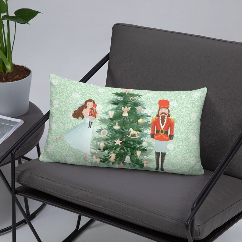 May include: A decorative pillow featuring a watercolor illustration of a ballerina, a nutcracker, and a Christmas tree with ornaments. The background is a light green with a white pattern.