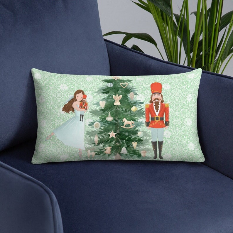 May include: A green and white throw pillow with a Christmas tree, a ballerina, and a nutcracker. The tree is decorated with ornaments, a star, and a rocking horse. The ballerina is wearing a blue dress and holding a doll. The nutcracker is wearing a red and blue uniform.