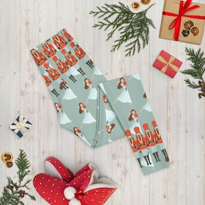 May include: A pair of blue leggings with a repeating pattern of a ballerina holding a gift and a nutcracker soldier. The leggings are on a white wood background with red and white slippers and a gift box.
