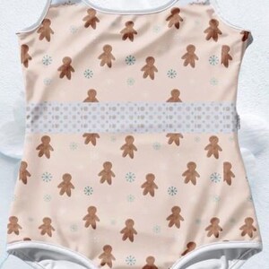 May include: A white one-piece swimsuit with a gingerbread man pattern on a light pink background. The swimsuit has a white trim and a white band with a polka dot pattern around the waist.