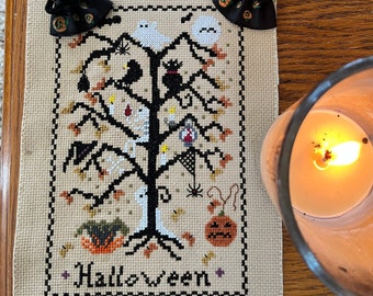 May include: A cross-stitch embroidery of a Halloween tree with a black cat, ghosts, bats, pumpkins, and spiders. The embroidery is framed with a black border and the word "Halloween" is stitched at the bottom.
