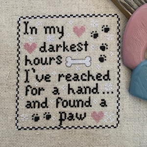 Puede incluir: Obra de arte de punto de cruz sobre tela beige con el texto "In my darkest hours I've reached for a hand and found a paw". Presenta corazones, huellas de patas y un hueso. El borde es blanco y negro. Un marco de madera y formas de madera pintadas están en el fondo.