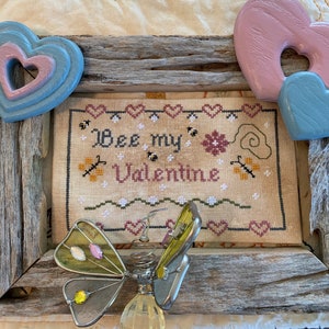 May include: A framed cross-stitch piece with the words "Bee my Valentine" and bee and heart motifs. The frame is rustic wood, and the image includes decorative wooden hearts and a metal butterfly.