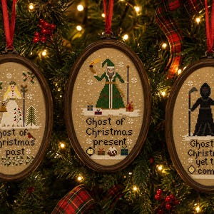 May include: Three oval-framed cross-stitch ornaments featuring figures from A Christmas Carol. The left reads Ghost of Christmas past, the center Ghost of Christmas present, and the right Ghost of Christmas yet to come, all with red ribbon hangers.