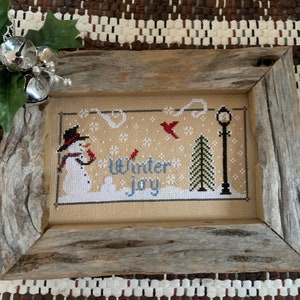 May include: A framed cross-stitch design featuring a snowman, a cardinal, a street lamp, and a pine tree. The design is on a beige background with the words "Winter Joy" in blue and white thread.