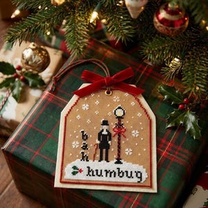 May include: A tan cross-stitch ornament shaped like a gift tag featuring a figure in a top hat standing by a lamppost, with the text "bah humbug" in black, white snowflakes, and a red velvet ribbon bow at the top, hanging by a green and red cord.