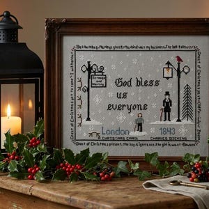 May include: A framed cross-stitch embroidery featuring the text God bless us everyone, London 1843, and Charles Dickens. The design includes street lamps, a figure, and falling snow on a gray background, all enclosed in a dark wooden frame.