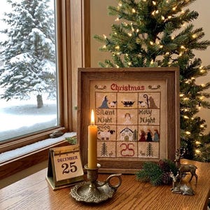 May include: A framed cross-stitch sampler featuring a grid of Christmas-themed motifs and the text "Christmas", "Silent Night", "Holy Night", and "25". A lit yellow taper candle in a brass holder and a metal desk calendar showing December 25.