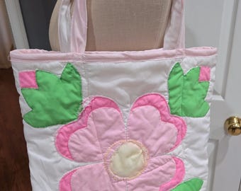 Könnte beinhalten: Eine weiße gesteppte Tragetasche mit einer rosa Blume und grünen Blättern. Die Tasche hat rosa Henkel.