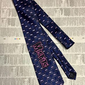 May include: A navy blue tie with a geometric pattern of small pink rectangles and squares. The tie features the word "MISFITS" in pink, stylized lettering. The tie is made of a smooth fabric and is displayed on a newspaper background.