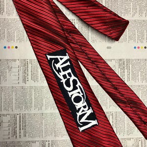 May include: A red and black striped necktie with the word "ALESTORM" in white lettering on a black rectangle. The tie is lying on a newspaper background.