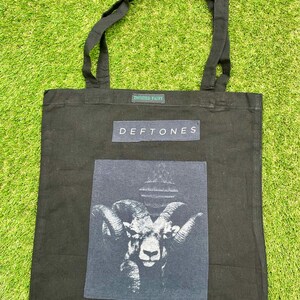May include: Black tote bag with the band name "Deftones" printed on a navy blue rectangle. The bag features a graphic of a ram's head. The text "Twisted Fairy" is printed above the band name.