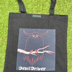 Puede incluir: Bolso tote negro con un gráfico de un búho con cuernos en una rama, en rojo y blanco, con el texto "DevilDriver" debajo. El bolso tiene dos asas negras y el texto "TWISTED FAIRY" en la parte superior.
