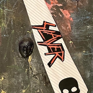 May include: A white striped tie with the word "SLAYER" in black and red lettering. A black skull graphic is at the bottom. The tie is set against a dark, textured background with abstract elements.