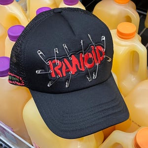 May include: Black mesh trucker hat with a red "RANCID" logo and safety pins. The hat is sitting on top of a stack of orange juice cartons.