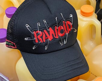 RANCID black trucker hat with band patch and safety pin details.