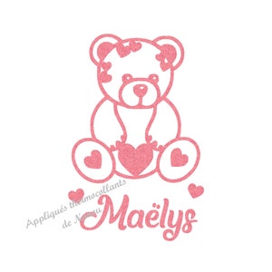 Può includere: Design di orsacchiotto rosa glitterato che tiene un cuore, con cuori sulla testa e sui piedi. Il nome "Maëlys" è scritto in corsivo sotto l'orso. È presente anche il testo "Appliqués thermocollants de Nanou".