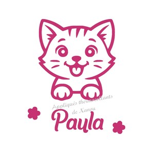 Peut inclure: Illustration d'un chat de dessin animé magenta avec un visage souriant et la langue tirée. Le texte "Paula" est en dessous du chat, avec les mots "Appliqués thermocollants de Nanou" au-dessus. Deux formes de fleurs magenta de chaque côté.