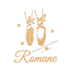 Peut inclure: Motif pailleté doré de chaussons de danse avec rubans et étoiles sur fond blanc. Le nom "Romane" est écrit en script doré pailleté. Texte : "Appliqués thermocollants de Nanou."
