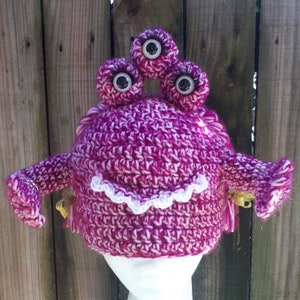 May include: A pink and white crocheted monster hat with three large eyes and two gold bells. The hat is on a white mannequin head.
