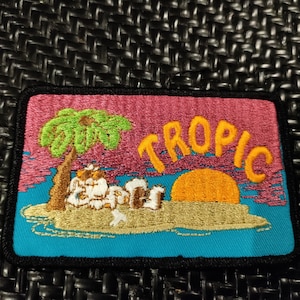 Peut inclure: Patch brodé avec un motif rose, bleu et jaune. Il représente un palmier, un soleil et le mot "TROPIC" dans une police de caractères stylisée.