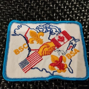 May include: Embroidered patch featuring a handshake between the flags of the United States and Canada, with a fleur-de-lis and a red flower. The patch is bordered in blue and has the letters "BSC" and "BSA" on it.