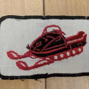 May include: Red and black embroidered patch of a snowmobile on a white background with a black border.