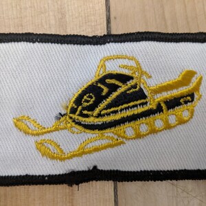 May include: A white embroidered patch with a black outline. The patch features a gold snowmobile with black details.