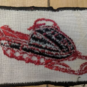 May include: A red and black embroidered patch of a snowmobile on a white background. The snowmobile is outlined in black and has red details. The patch is surrounded by a black border.