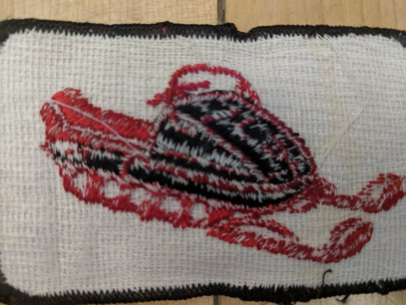 May include: A red and black embroidered patch of a snowmobile on a white background. The snowmobile is outlined in black and has red details. The patch is surrounded by a black border.