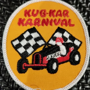 Peut inclure: Un écusson brodé jaune et blanc avec le texte "KUB-KAR KARnival" en rouge. L'écusson représente un karting rouge avec des roues noires et un drapeau à damier.