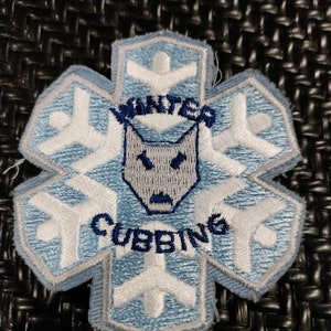 Peut inclure: Un écusson brodé bleu et blanc avec un motif de flocon de neige. Le centre du flocon de neige présente une tête de loup avec les mots "Winter Cubbing" en dessous.