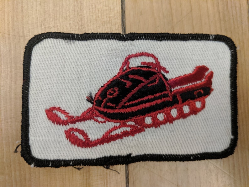 May include: Red and black embroidered patch of a snowmobile on a white background with a black border.