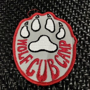 May include: A grey and red embroidered patch with a white paw print in the centre. The text "Wolf Cub Camp" is written around the paw print.