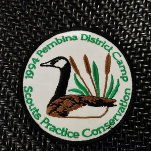 Peut inclure: Un écusson rond blanc avec des broderies vertes et noires. Il représente une oie aux plumes brunes et noires, des roseaux verts et le texte "1994 Pembina District Camp Scouts Practice Conservation".
