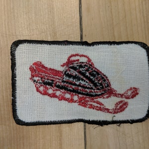 May include: A red and black embroidered patch of a snowmobile on a white background with a black border.