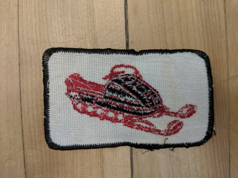 May include: A red and black embroidered patch of a snowmobile on a white background with a black border.