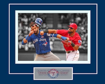 Op de afbeelding: Een actiefoto van een honkbalwedstrijd met twee spelers, een in een blauw shirt met de tekst "Blue Jays" en de ander in een rood shirt met de tekst "T". De speler in het blauwe shirt zwaait met zijn knuppel, terwijl de speler in het rode shirt probeert de bal te vangen. De tekst "SMACK DOWN IN TEXAS" is zichtbaar onderaan de afbeelding.