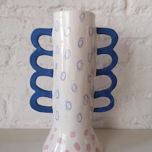 Puede incluir: Un jarrón de cerámica blanco con asas azules y una base con estampado rosa y blanco. El jarrón tiene un diseño único y abstracto con círculos azules sobre el cuerpo blanco.